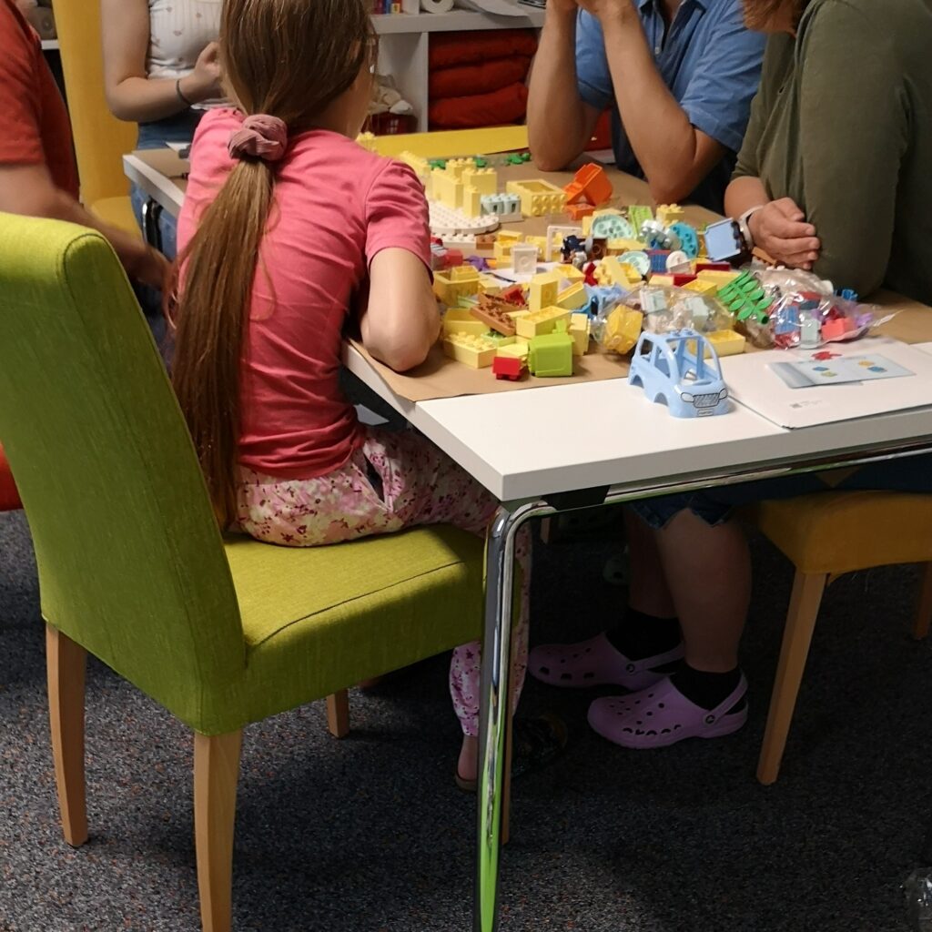 Ein Kind sitzt an einem Tisch mit vier Erwachsenen. Auf dem Tisch sind verschiedene Legosteine und ein Teil von einem Spielzeugauto erkennbar. 