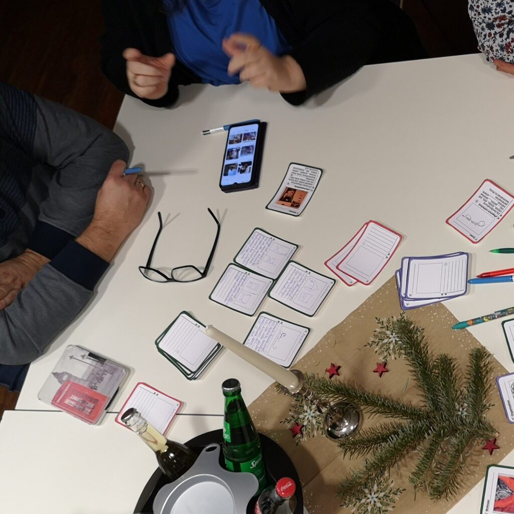 Auf einem Tisch liegen kleine Zettel. Links liegen ein Handy und eine Uhr. Unten rechts ist Tischdekoration mit Tannenzweigen und Sternen und Getränkeflaschen erkennbar. Es sitzen drei Personen an dem Tisch. 